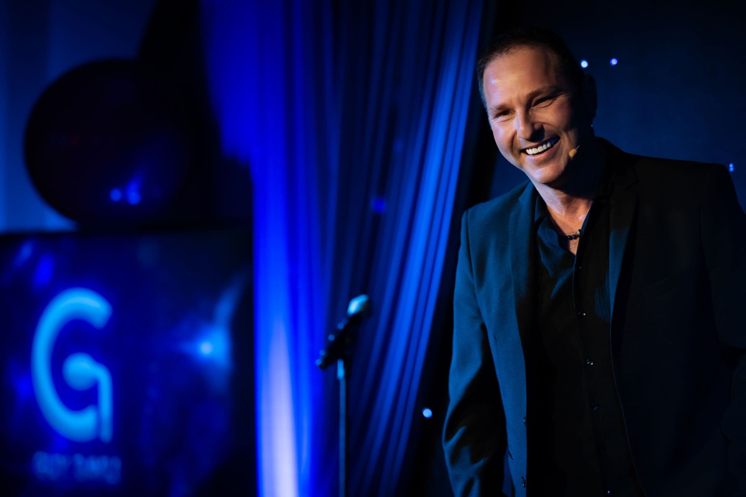 Guy Bavli smiling at the microphone on a blue-lit theatre stage